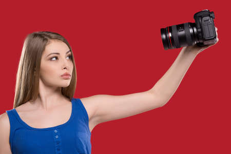Women with camera. Confidence young women photographing herself while standing isolated on red backgroundの写真素材
