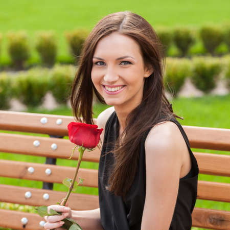 Girl with flower. Beautiful young woman holding red rose and smiling while sitting on the benchの写真素材