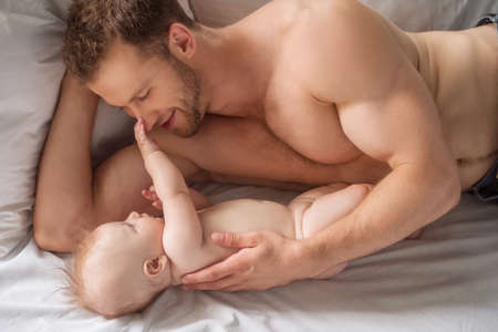 Man with little baby. Young muscular man and little baby lying on the bed together and looking at each otherの写真素材