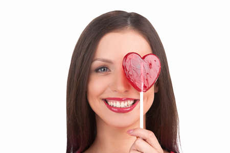 Girl with lollipop. Portrait of beautiful young woman holding heart shape lollipop in front of her eye while isolated on whiteの写真素材