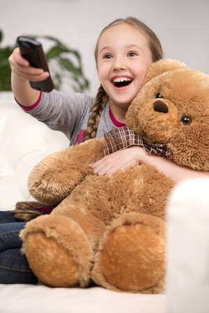 Cute girl at home. Cheerful little girl watching TV and holding toy bearの写真素材