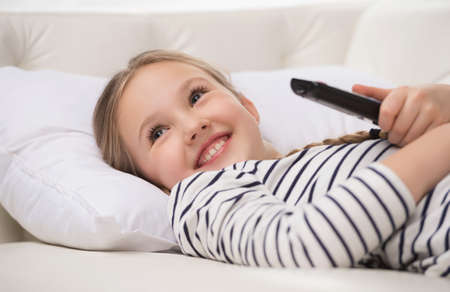 Girl watching TV. Cheerful little girl holding remote control and smilingの写真素材