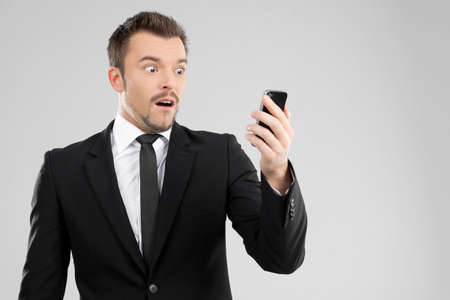 Shocked businessman. Surprised young man in formalwear looking at mobile phoneの写真素材