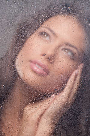 Girl dreaming. Beautiful young woman looking through wet window and smilingの写真素材