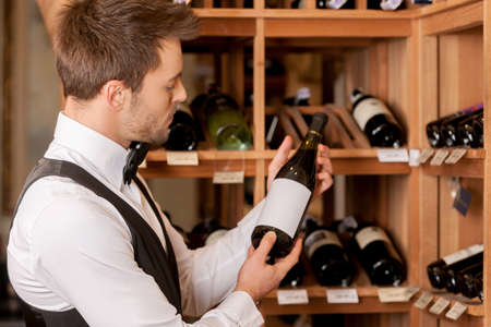 Confident sommelier. Thoughtful young sommelier holding a wine bottle and examining itの写真素材