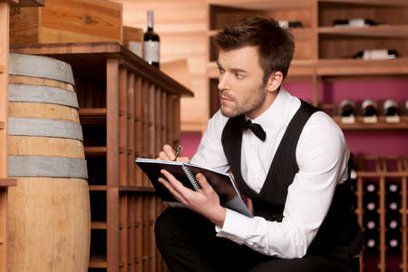 Confident sommelier  Confident young sommelier writing something at his note pad and looking at wine barrelの写真素材