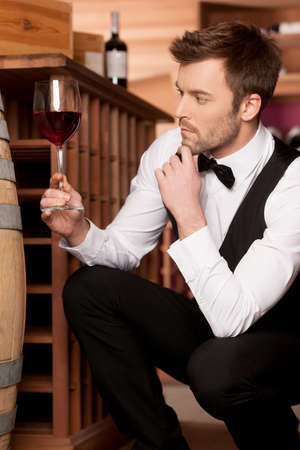 Confident sommelier  Thoughtful young sommelier holding a glass of wine while sitting near wine barrelの写真素材