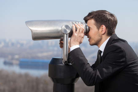 Looking for solutions. Side view of cheerful young men in formalwear looking through stationary binoculars and smilingの写真素材