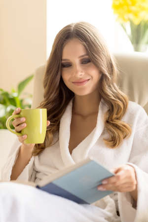 Day off at home. Beautiful young woman in bathrobe drinking coffee and reading bookの写真素材