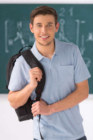Happy student. Handsome young men holding backpack and smiling while standing in front of the blackboardの写真素材
