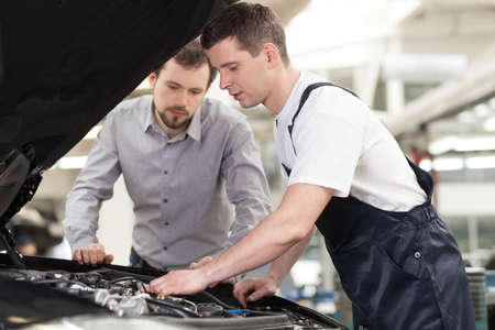 Examining a car. Confident auto mechanic examining car engine while customer looking at itの写真素材