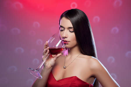 Drinking red wine  Beautiful young woman in red dress holding a glass with wine while standing isolated on colored backgroundの写真素材