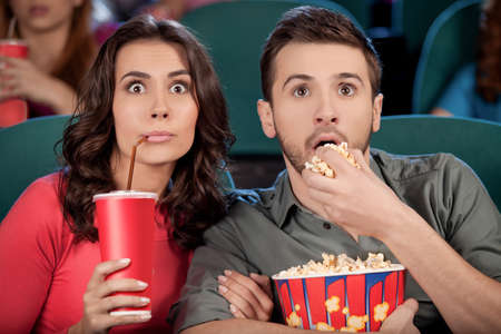 Exciting movie. Shocked young couple eating popcorn and drinking soda while watching movie at the cinemaの写真素材
