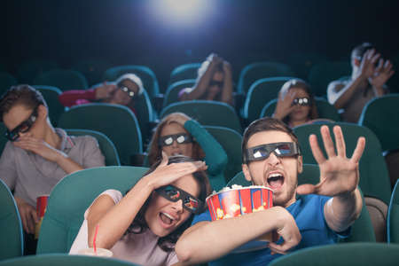 Watching three-dimensional movie. Terrified people in three-dimensional glasses watching movie at the cinema and gesturingの写真素材