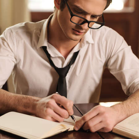Capturing his thoughts. Handsome young author sitting at the table and writing something in his sketchpadの写真素材