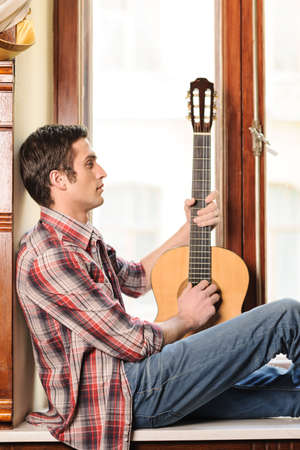 Looking for inspiration. Handsome young men sitting on the windowsill and holding an acoustic guitar in his handの写真素材