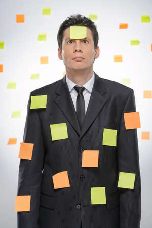 Mired in work. Frustrated businessman covered with the adhesive notes standing against the wall with sticky notes on itの写真素材