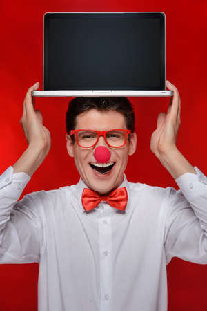 Holding laptop on his head  Cheerful man with clown holding a laptop on his head while standing isolated on redの写真素材