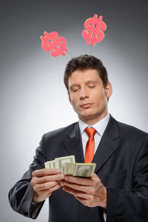 Money-maker  Funny businessman with a dollar hornes on the head counting money against grey backgroundの写真素材