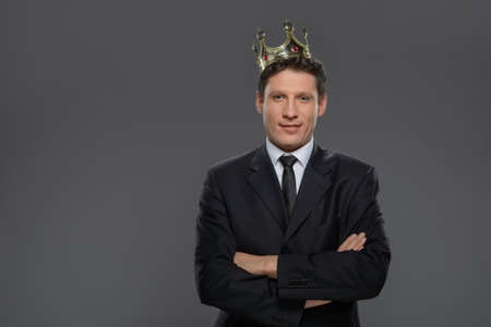 Business king. Confident businessman in crown standing isolated on gray with his arms crossed