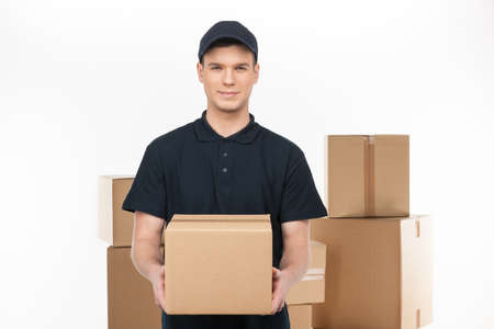 Confident deliveryman.  Cheerful young deliveryman holding a cardboard box while standing in front of the box stackの写真素材