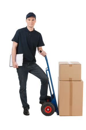 Box moving. Cheerful young deliveryman leaning on the cart with boxes on it while isolated on whiteの写真素材