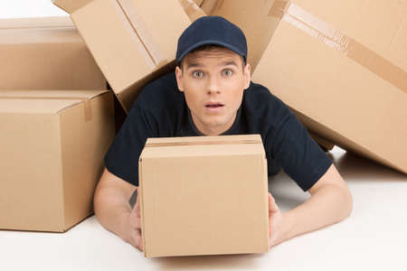 Be careful with fragile. Deliveryman lying covered with a stack of cardboard boxes and looking at cameraの写真素材