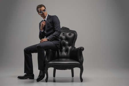 Looking just perfect. Full length of confident young businessmen in sunglasses sitting on the vintage chair while isolated on greyの写真素材
