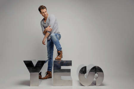 Just say ï¿½esï¿½ Handsome young men stepping on the metal letter and looking away while isolated on greyの写真素材
