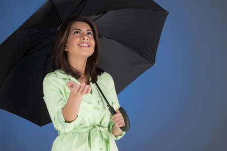 Rainy day. Attractive young women holding an umbrella and looking up while isolated on blue backgroundの写真素材