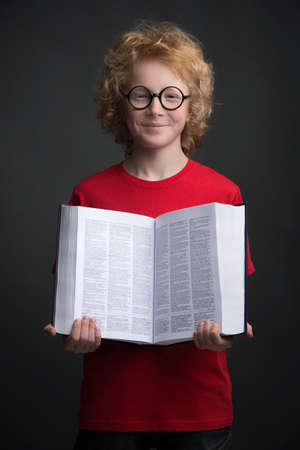Smart child. Cheerful red hair boy in glasses hoilding a book while standing isolated on blackの写真素材