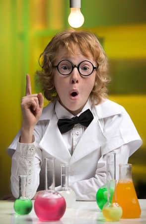 I have an idea! Happy little boy in lab coat gesturing while sitting at the table with laboratory equipment on itの写真素材