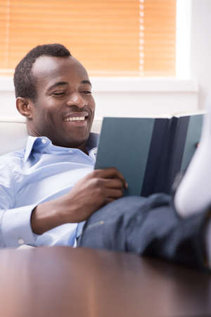 Reading an exciting book. Cheerful African descent reading a book while sitting on the couch.の写真素材