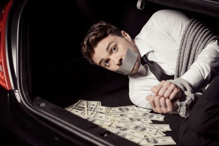 Kidnapped man. Tied up businessman lying in the car trunk full of paper currency and looking at cameraの写真素材