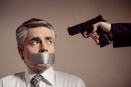 Victim of kidnapping. Shocked mature businessman looking at the handgun aimed on him by kidnapperの写真素材