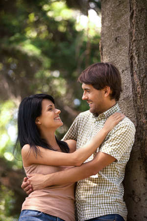 Beautiful young couple hugging and looking at each other while leaning on the tree in a parkの写真素材
