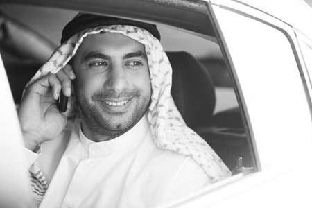 Cheerful Arab businessman. Black and white image of smiling Arab businessman sitting on the back seat of car and talking on the mobile phoneの写真素材