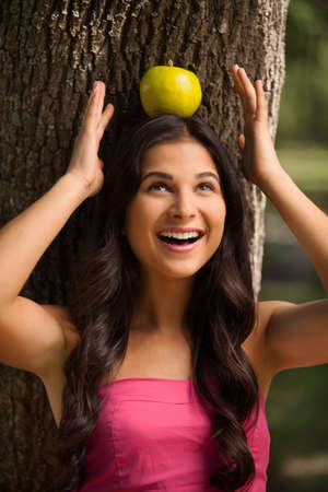 Apple on her head. Beautiful young woman standing back to the tree and holding apple on her headの写真素材