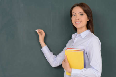 Confident teacher. Cheerful young teacher standing near blackboard and smiling at cameraの写真素材