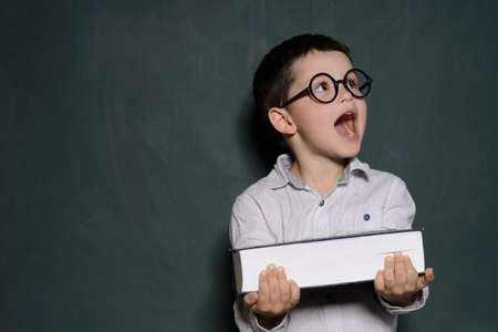 Happy schoolboy. Cheerful little schoolboy in glasses holding a book and laughing while standing in front of blackboardの写真素材