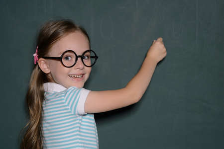 Happy schoolgirl. Cheerful little schoolgirl in glasses standing near the blackboard and looking over shoulderの写真素材