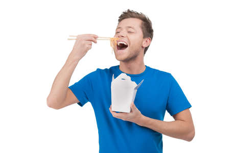 Man eating noodles. Cheerful young man eating rice from the food box while standing isolated on whiteの写真素材