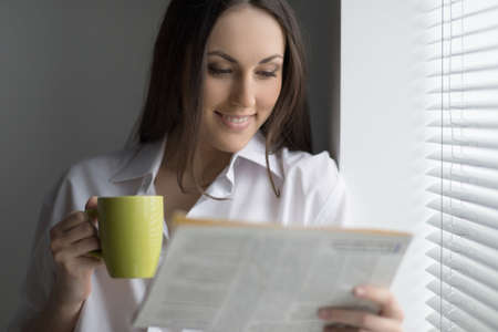 Hot coffee and fresh news. Beautiful young woman reading newspaper and drinking coffee