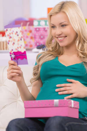 Happy pregnant woman. Cheerful young pregnant woman looking at the greeting card and holding a gift...