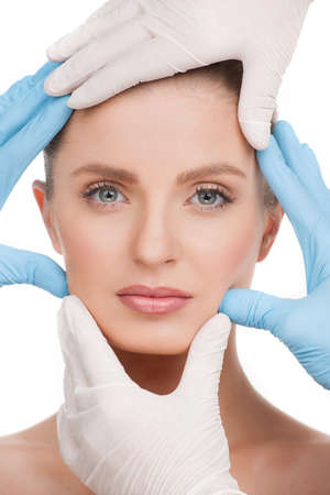 Face examining. Portrait of beautiful young woman looking at camera while many hands in gloves examining her faceの写真素材