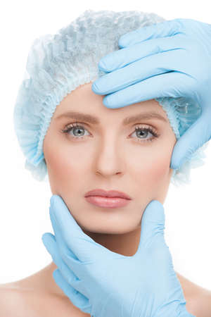 Examining face before surgery. Portrait of beautiful young woman in medical headwear looking at camera while hands in gloves examining her face isolated on whiteの写真素材