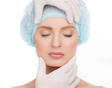 Examining face before plastic surgery. Portrait of beautiful young woman in medical headwear keeping eyes closed while hands in gloves examining her face isolated on whiteの写真素材