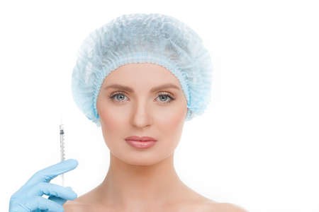 Syringe near face. Portrait of beautiful young woman in medical headwear looking at camera while hand in glove holding a syringe near her face isolated on whiteの写真素材