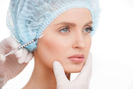 Medical injection. Portrait of beautiful young woman in medical headwear looking away while hand in glove making an injection in her face isolated on whiteの写真素材