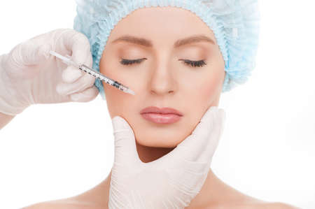 Medical injection. Portrait of beautiful young woman in medical headwear keeping eyes closed while doctors hands in gloves making an injection in her face isolated on whiteの写真素材